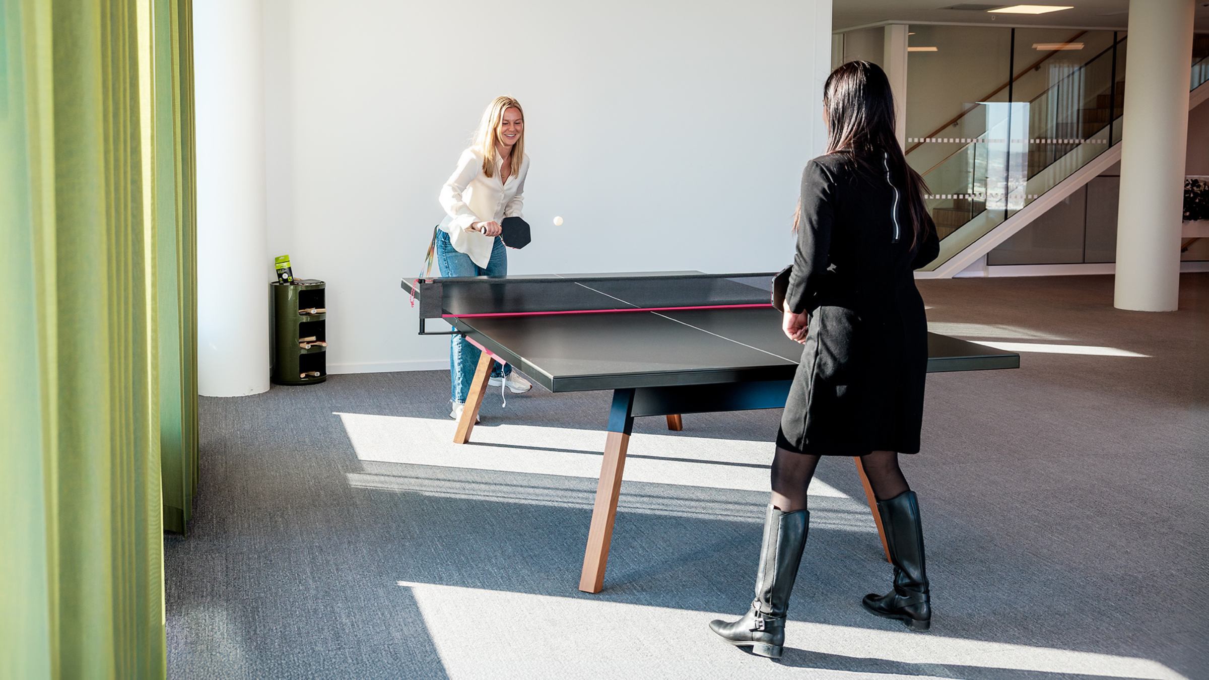 Office Ping Pong Match: Women Enjoying a Friendly Game Två kvinnor spelar bordtennis i en modern kontorsmiljö med naturligt ljus som strömmar in.