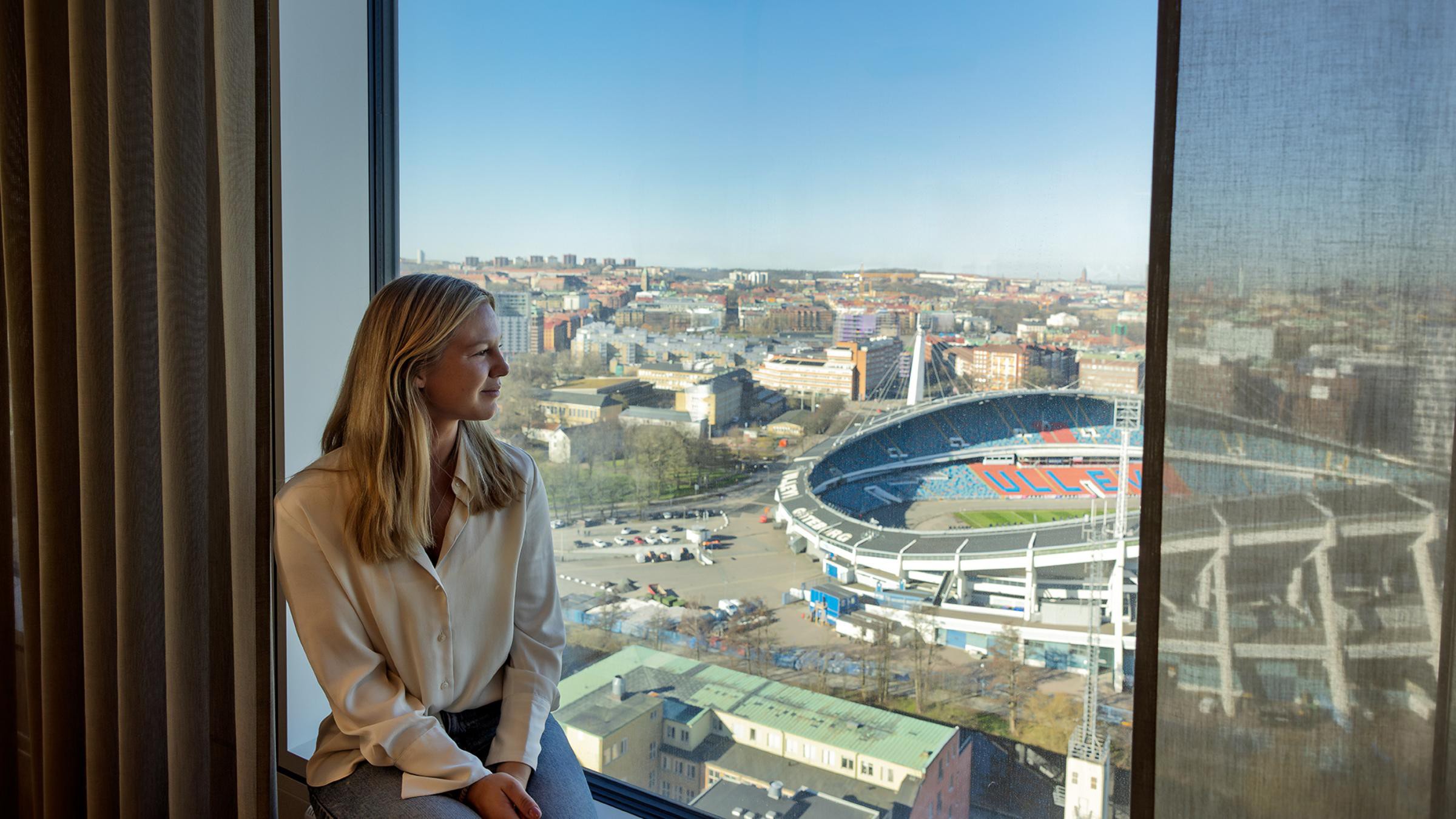 Stadsbildsutsikt från höghus nära stadion En person i en vit blus sitter vid ett fönster med utsikt över en stor stadion och stadsbild.