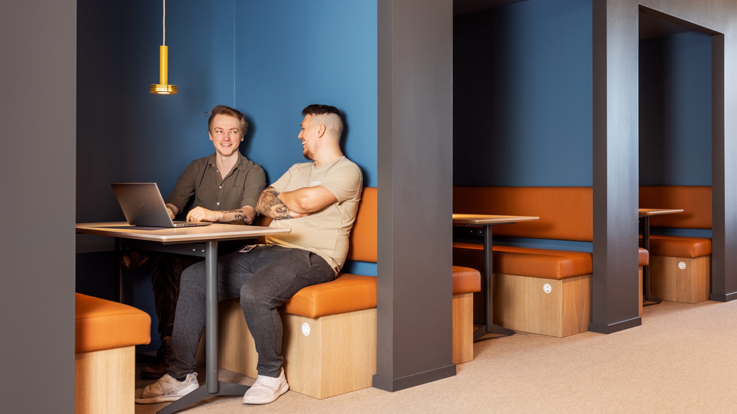 Modern samarbetsarbetsplats med stilfulla båsplatser Två personer sitter vid ett bord i en modern, blåväggig bås med orange sittplatser och en laptop.