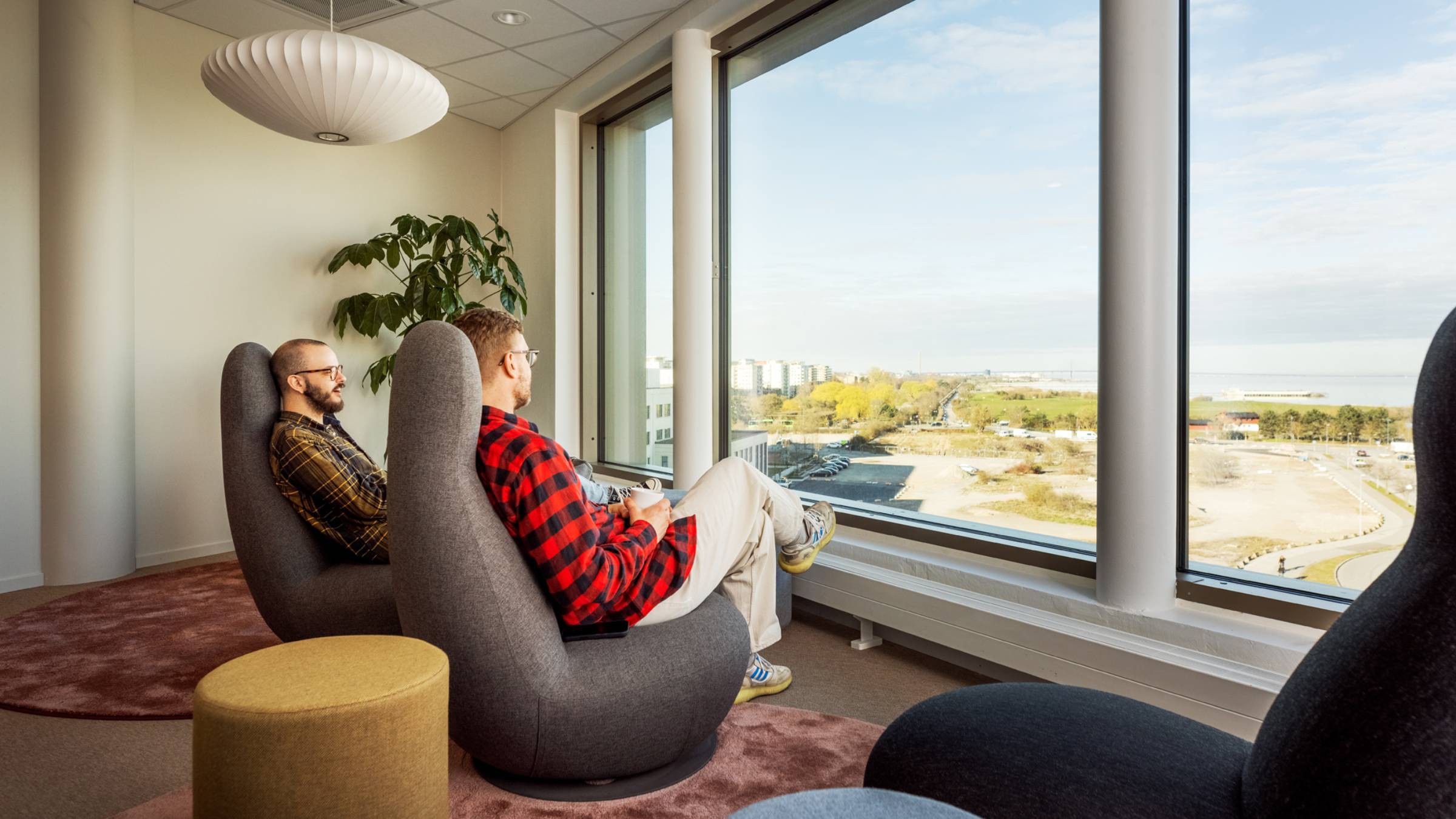 Modern kontorslounge med naturskön stadsutsikt Två personer kopplar av i moderna stolar vid ett stort fönster med utsikt över ett naturskönt stadslandskap.