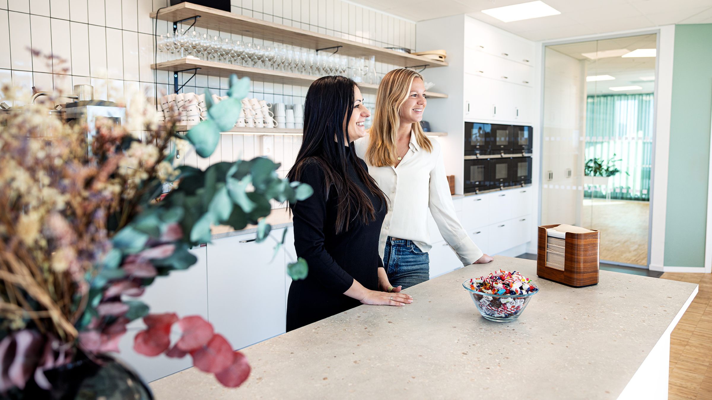 Modern kontorskök med stilfull inredning och godisskål Två kvinnor i ett modernt kök med hyllor, en godisskål och dekorativa växter.
