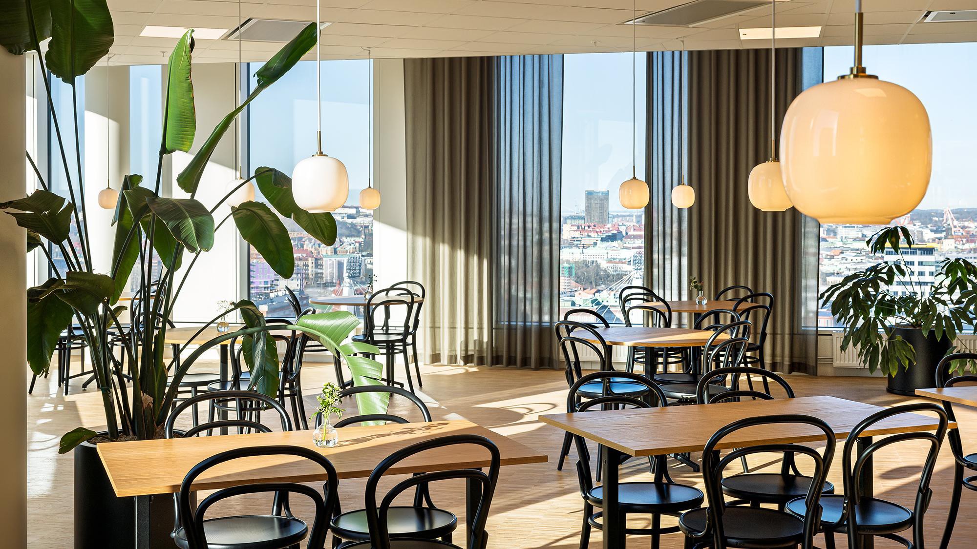 Modern caféinteriör med stadsutsikt och stilfull belysning Modern matplats med träbord, svarta stolar, stora fönster och hängande pendellampor.