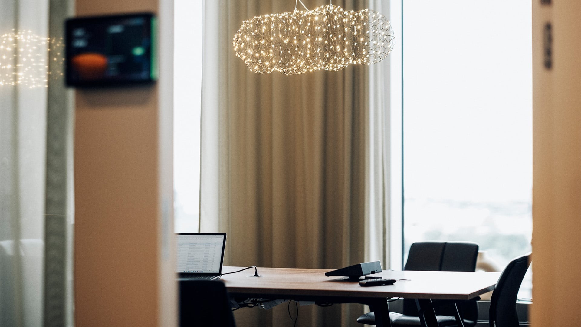 Modern konferensrum med ett träbord, laptop, stolar och en dekorativ belysningsarmatur.