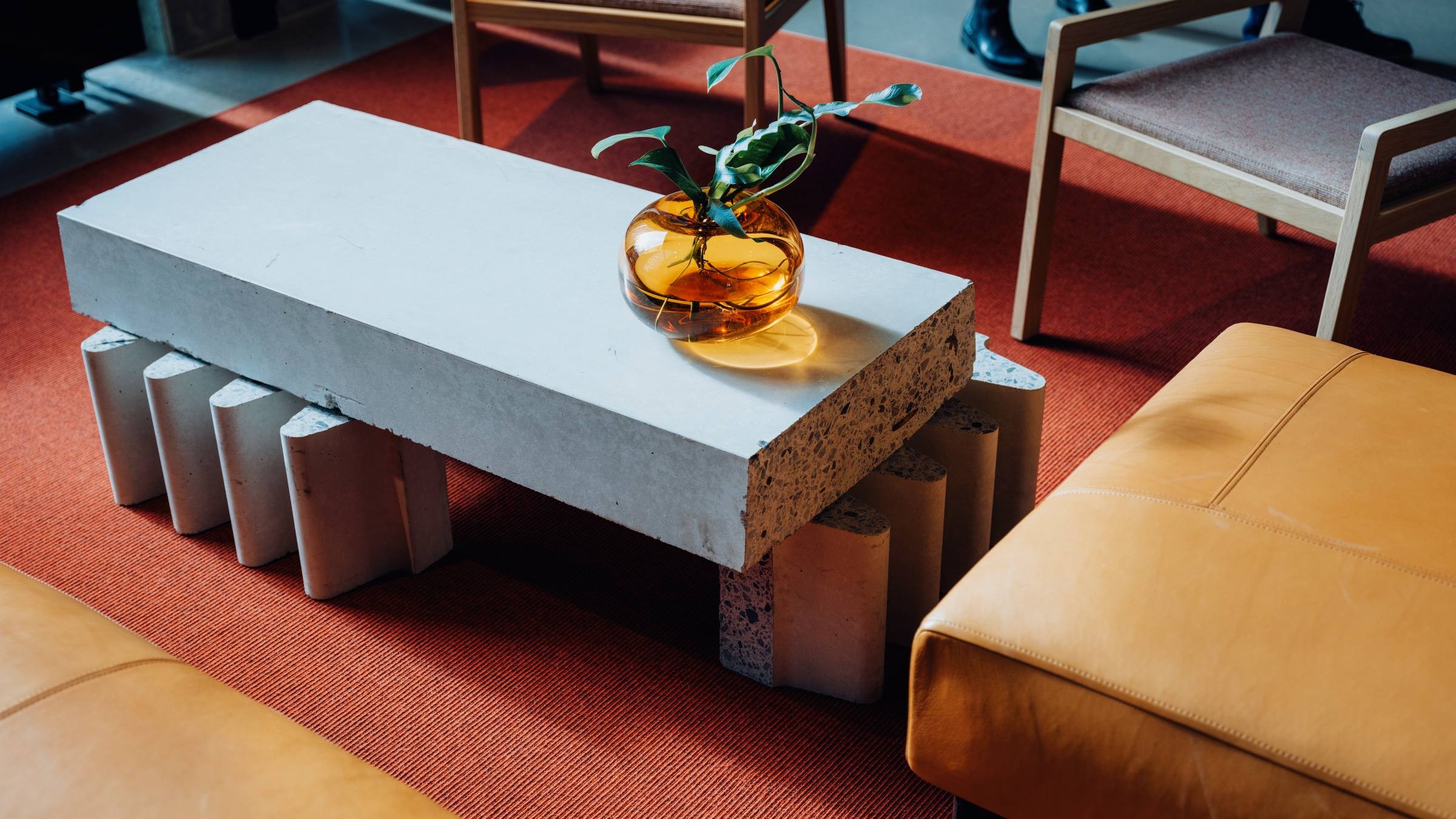 Modern vardagsrum med unik kaffebord i betong Modern betongsoffbord med dekorativ bärnstensvas på en röd matta, omgiven av sittplatser.