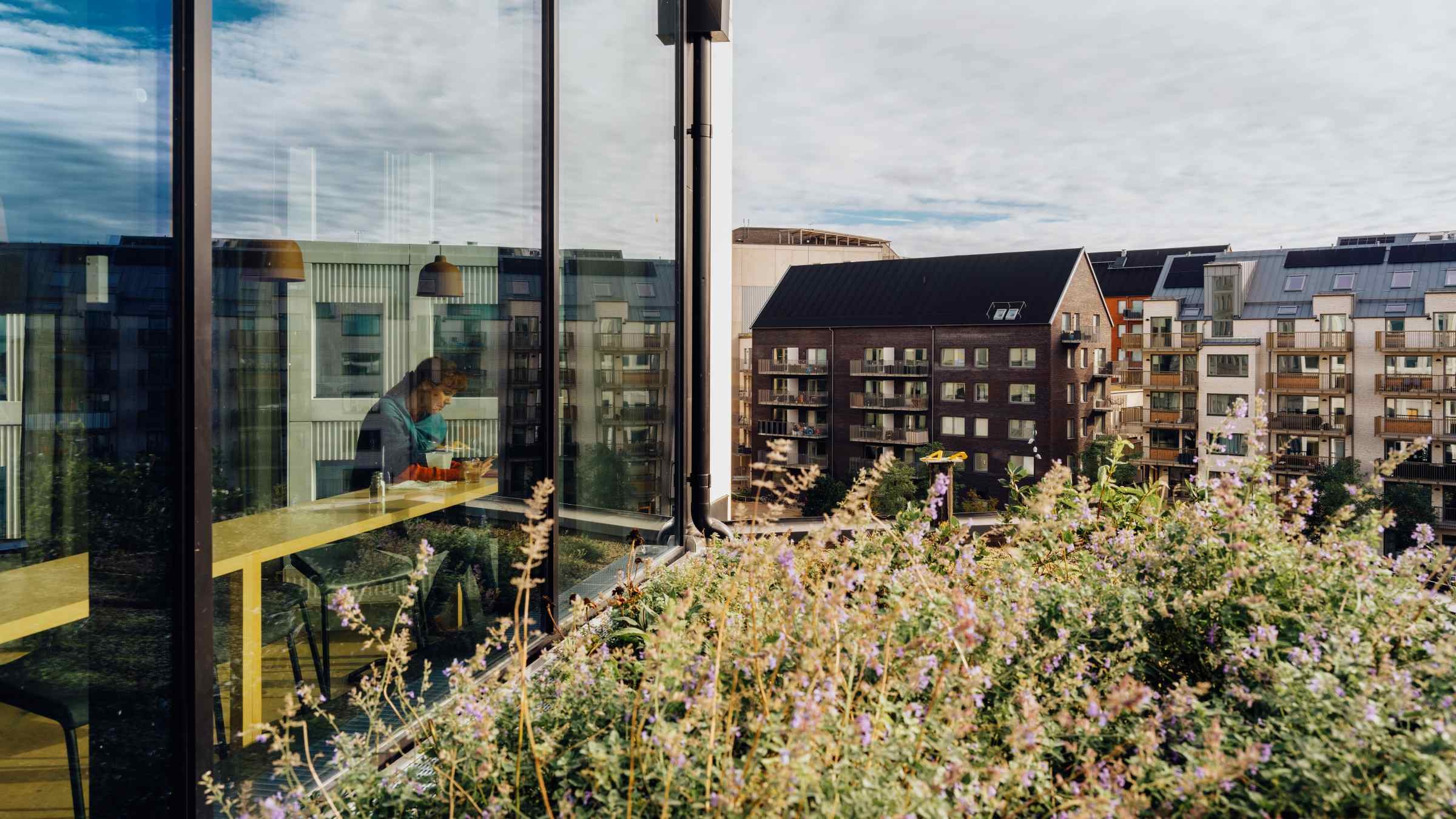 Modern Urban Living: Takutsikt med Grönska En man arbetar vid ett skrivbord inne i en modern glasbyggnad, omgiven av urbana lägenheter och växter.