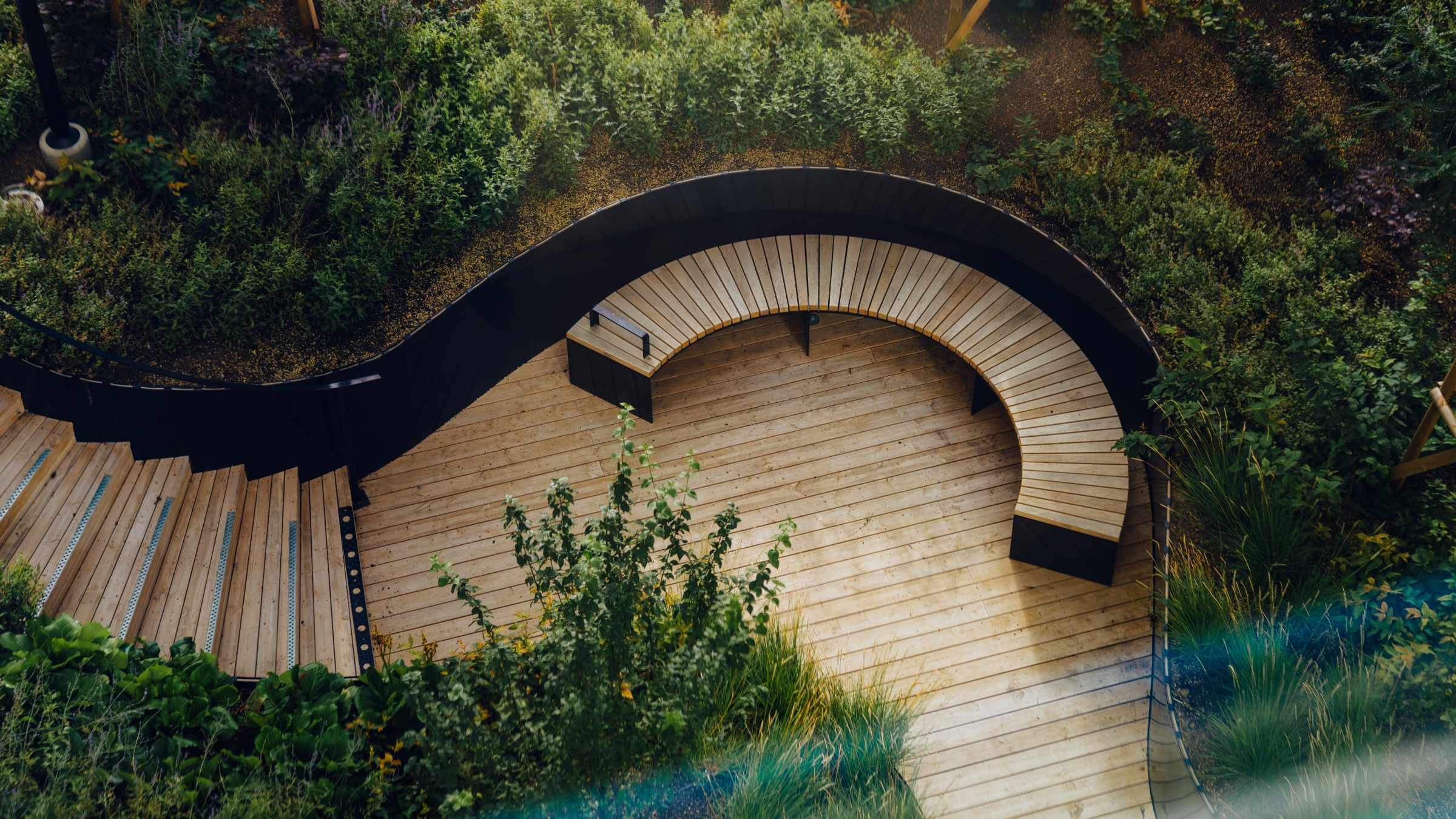 Böjd träaltan i frodig trädgårdslandskapsdesign Böjd träpromenad med integrerade sittplatser omgiven av frodig grön trädgårdslövverk.