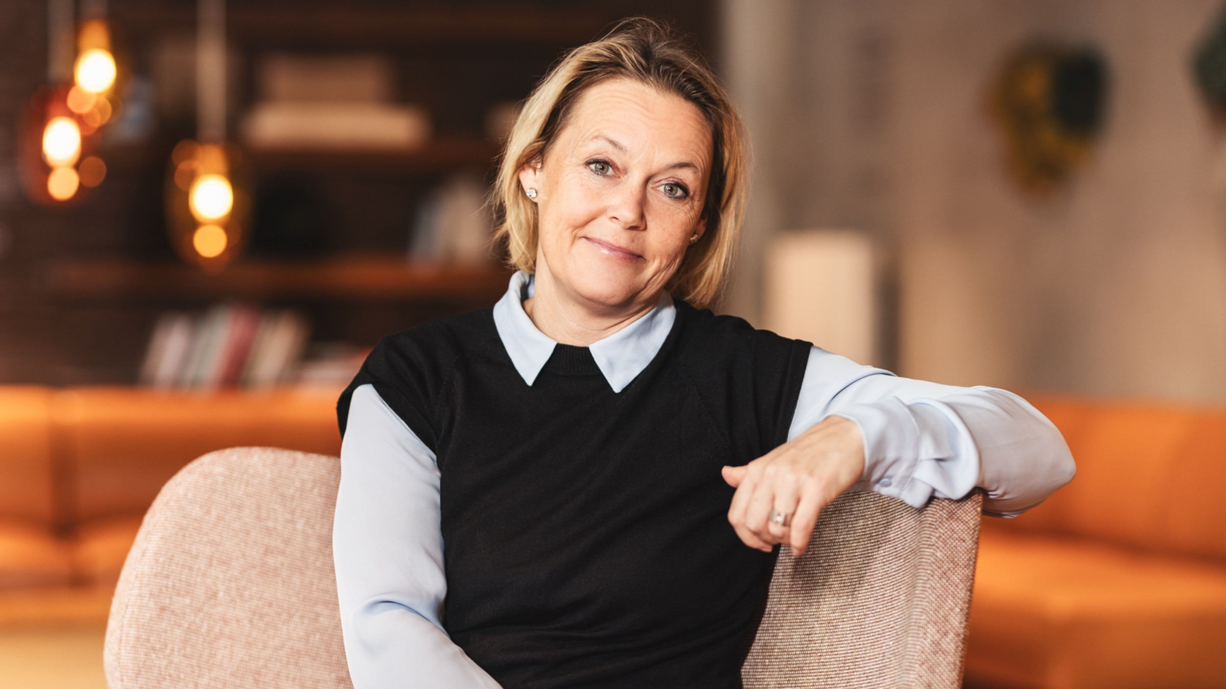 Professionell kvinna som kopplar av i modern mysig kontorsmiljö En person i en stilfull svart och blå outfit sitter bekvämt i ett modernt, varmt belyst rum.