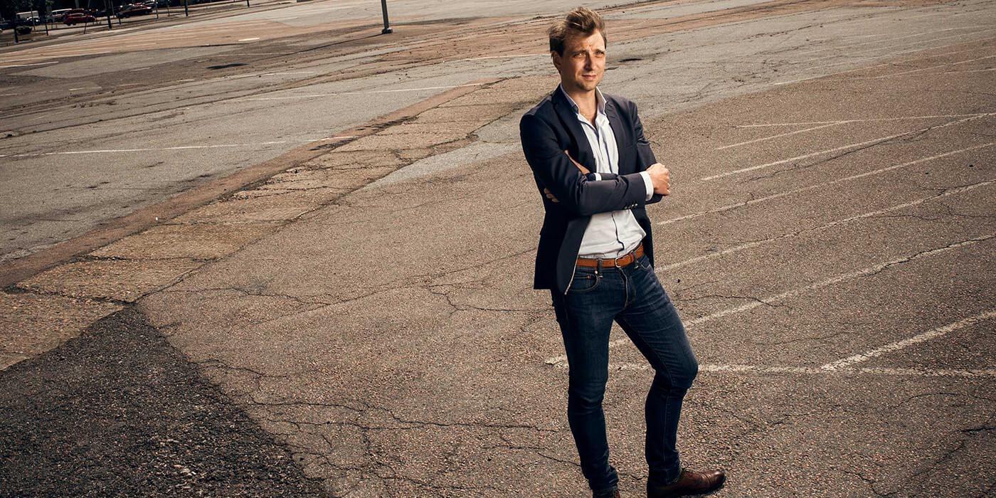 En man i en stilig blazer och jeans står självsäkert på en sprucken, tom parkeringsplats.