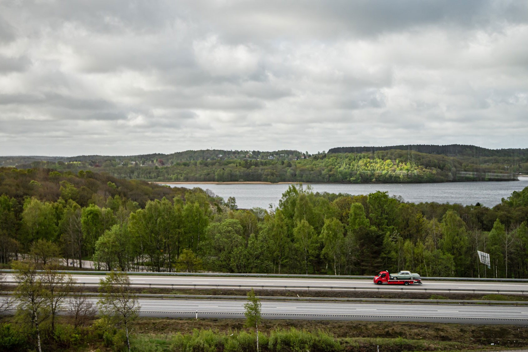En röd bärgningsbil kör på en motorväg, omgiven av frodiga gröna träd och en fridfull utsikt över en sjö.