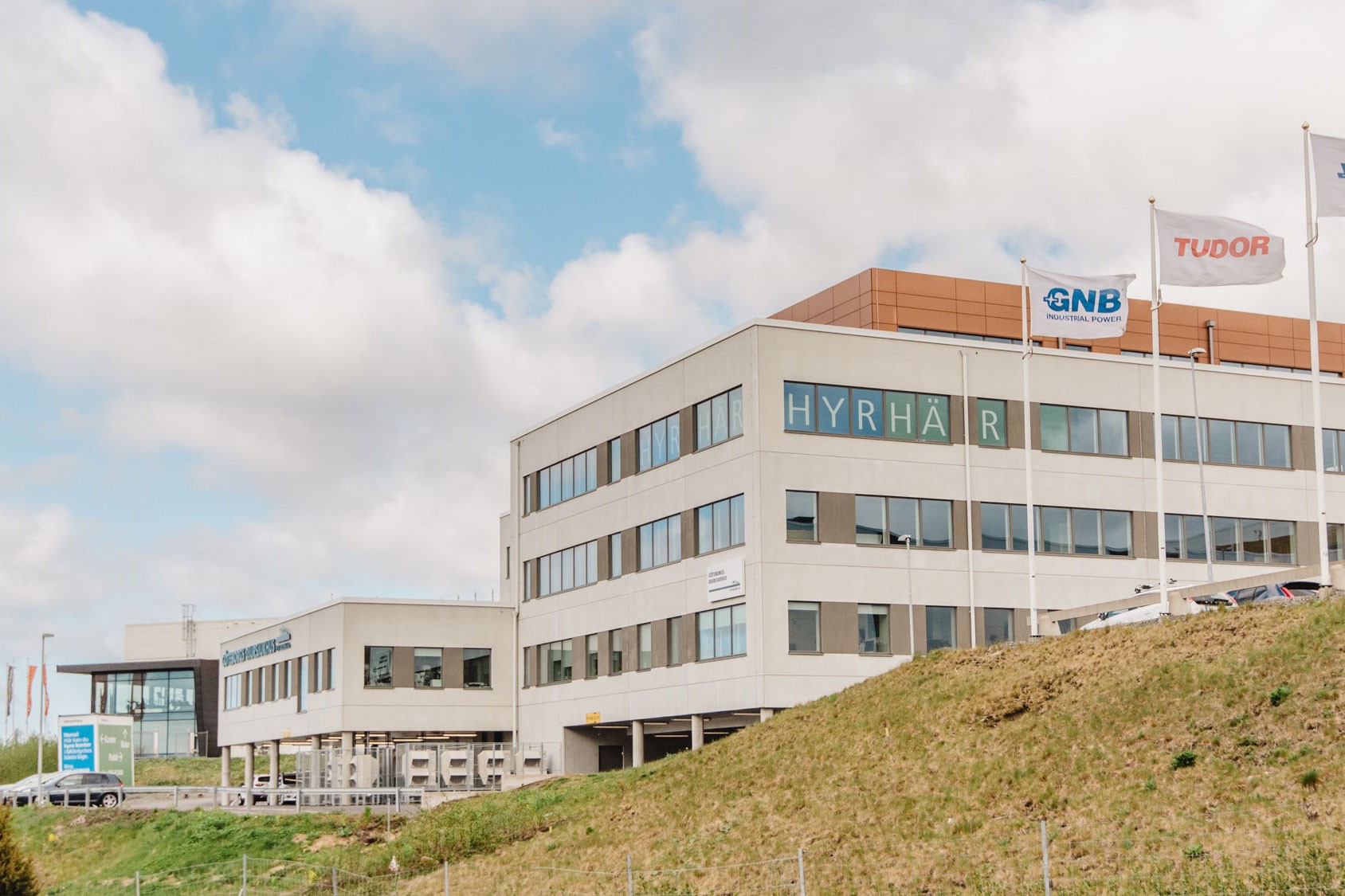 Modern kontorsbyggnad med "HYRHÄR"-skylt, omgiven av flaggor och grönskande landskap.