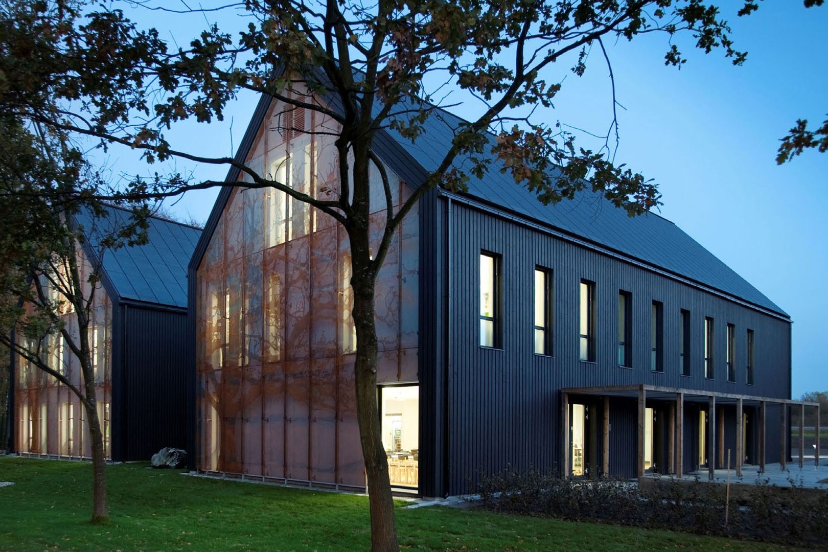 Modern byggnad med mörk exteriör, stora fönster och upplyst interiör, omgiven av träd.