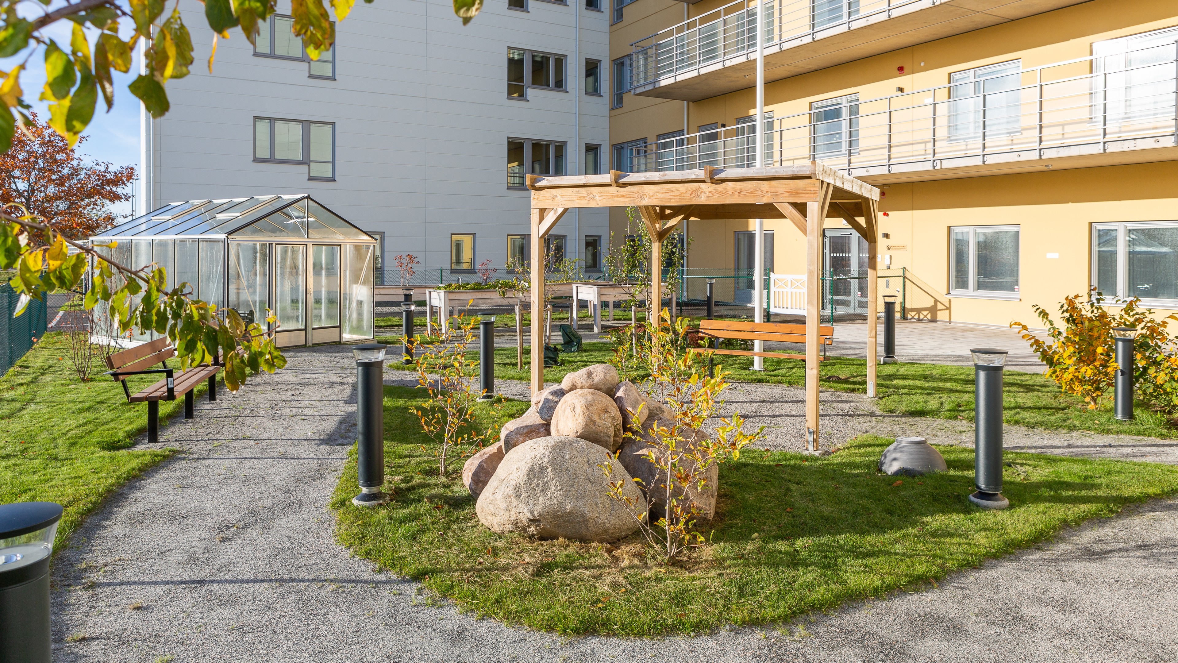 Modern utomhusträdgård med bänkar, pergola och ett växthus nära en lägenhet.