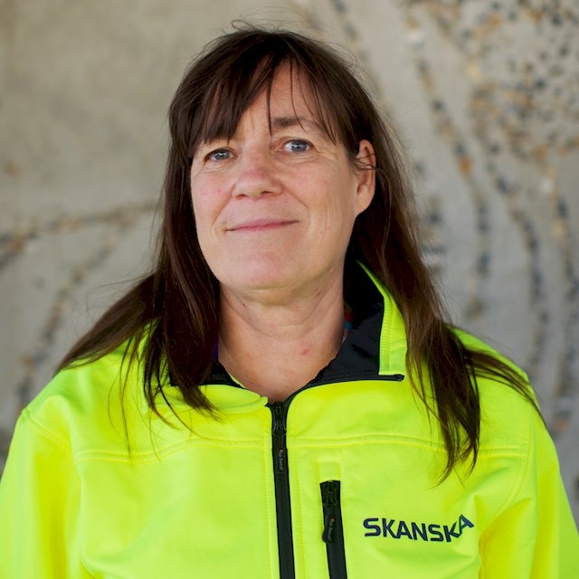 Anja Skans Person som bär en gul Skanska-jacka stående mot en texturerad väggbakgrund.