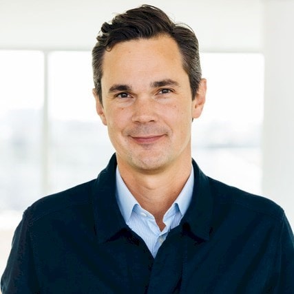Arvid Norman Person in navy blue jacket and light blue shirt against a bright window background.