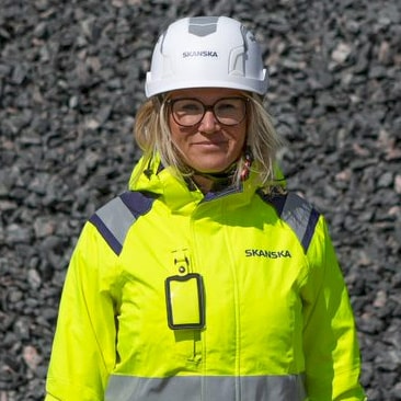 Barbro Brattström Grujovic En Skanska-arbetare i en ljusgul säkerhetsjacka och hjälm står framför en stenig bakgrund.