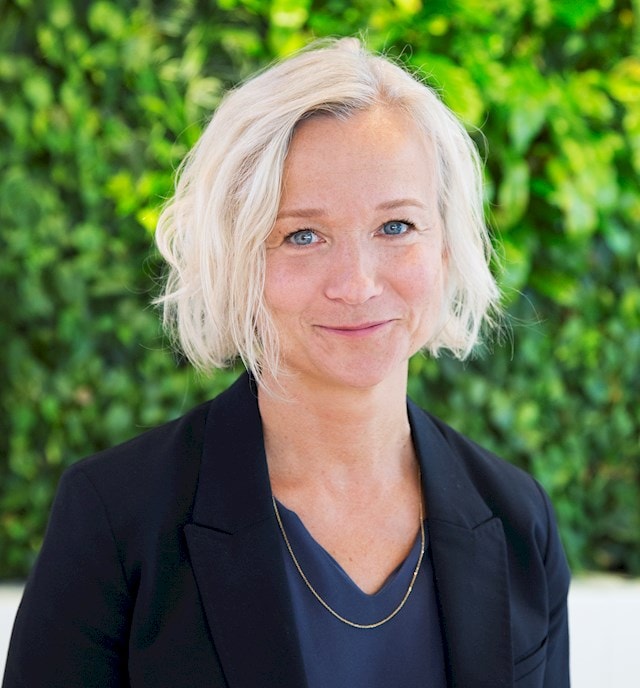 Camilla A Wieslander Person with short blond hair wearing a dark blazer and necklace outdoors.