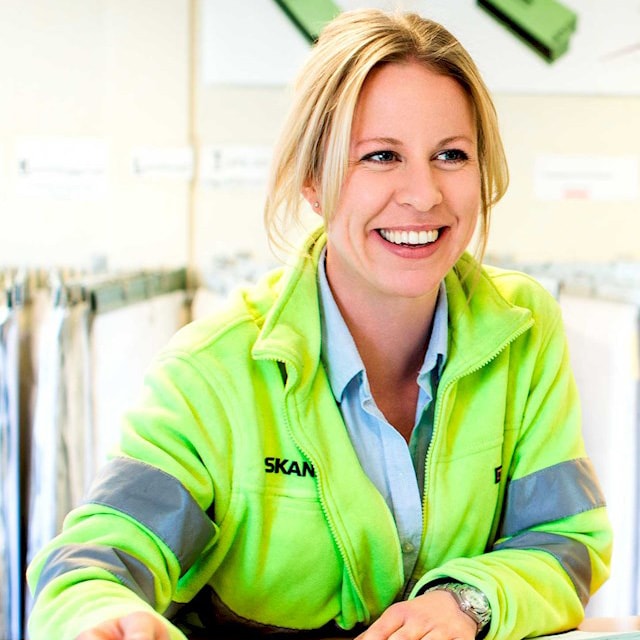 Caroline Apelqvist En person i en neongul Skanska-jacka arbetar vid ett skrivbord i en professionell miljö.