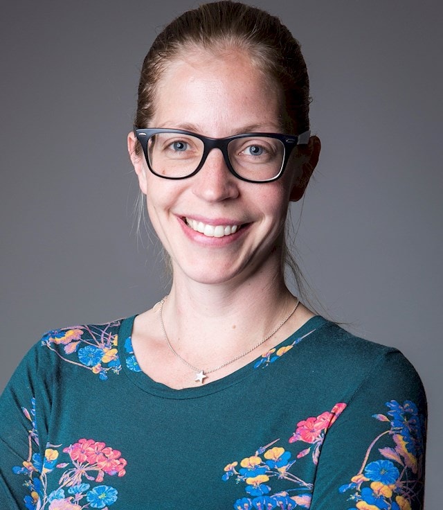 Evelina Källström En person som bär en blommönstrad teal skjorta och glasögon står mot en grå bakgrund.