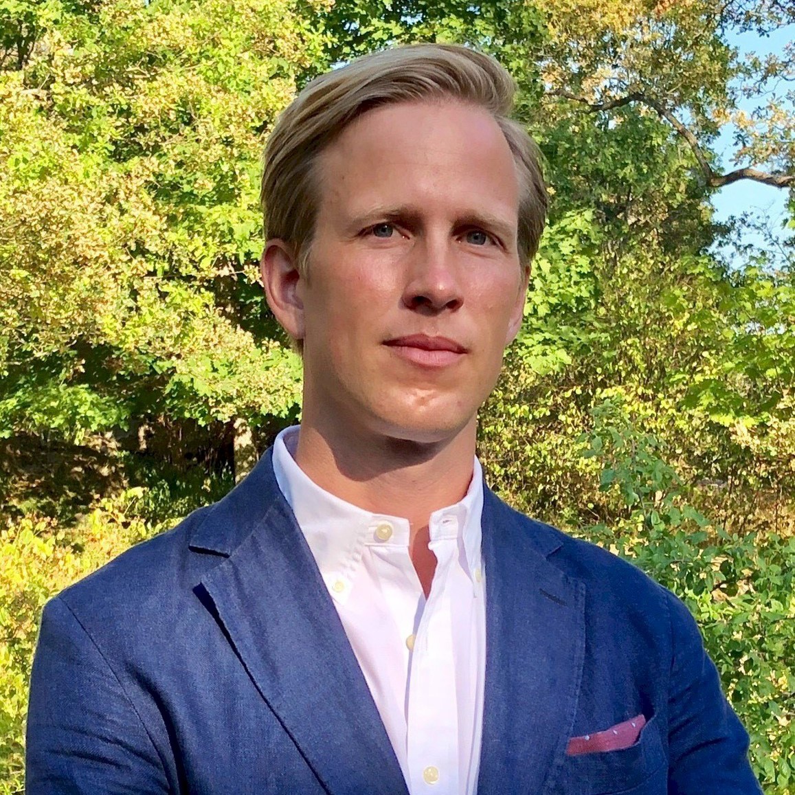 Gustav Ossmer Ung person i blå blazer och vit skjorta som står utomhus med grön träd bakgrund.