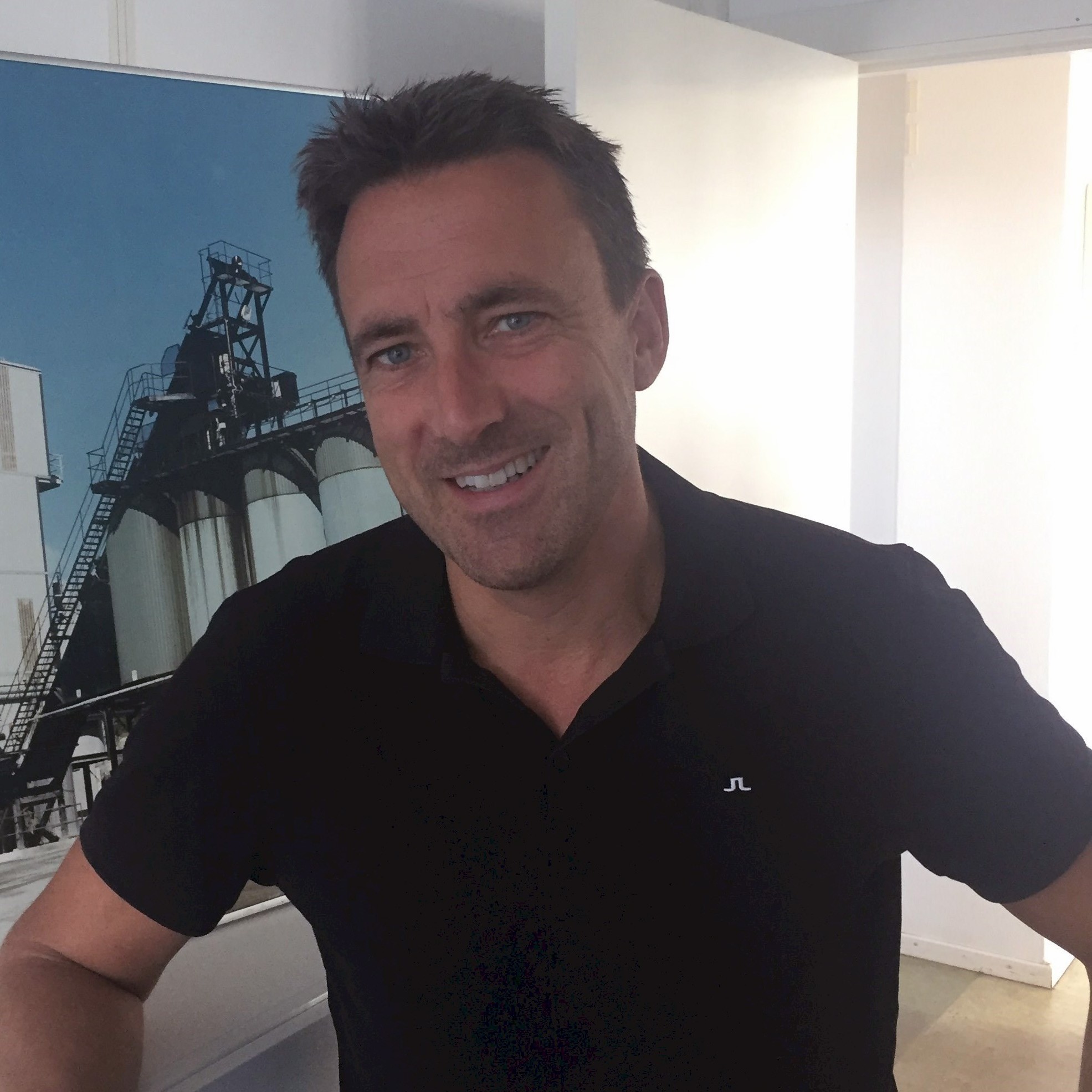 Henrik Sjöholm En person i en svart skjorta står nära ett stort industriellt foto som föreställer silos och metallstrukturer.
