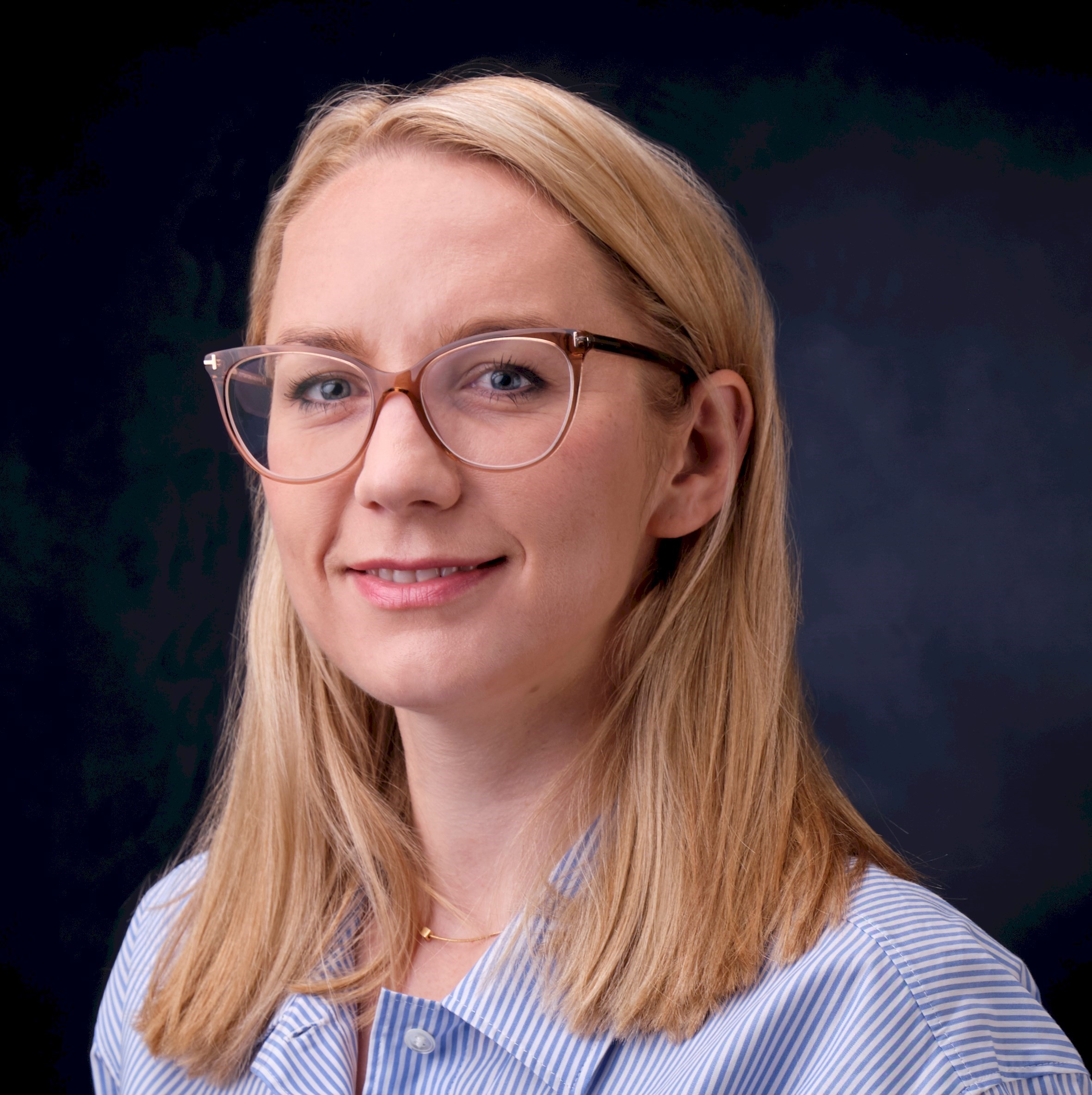 Izabela Kurkowska Blond person med glasögon som bär en blå randig skjorta mot en mörk bakgrund.