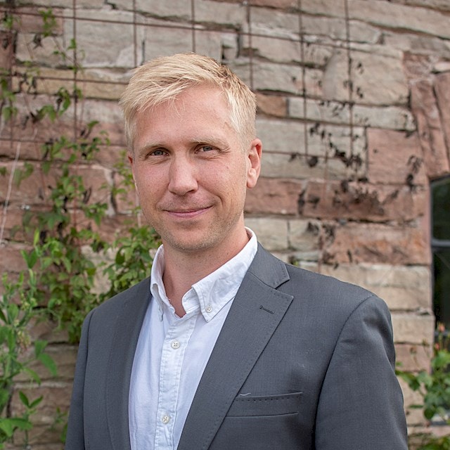 Jacob Olsson-Wallentin Man i grå kostym och vit skjorta står mot en stenmur med grönska.