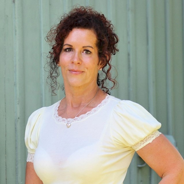 Jeanette Almberg En kvinna med lockigt hår bär en vit blus med spetsdetaljer, stående mot en grön bakgrund.