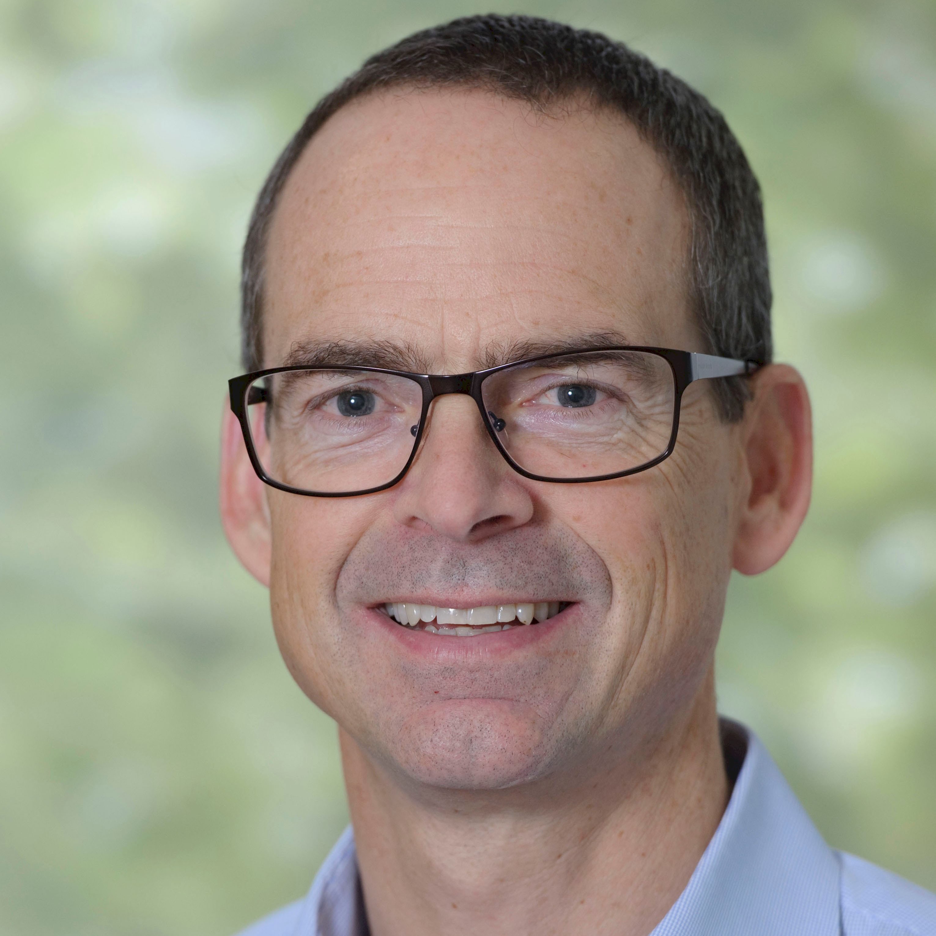 John Dingle A person with glasses and a blue collared shirt stands against a green blurry background.