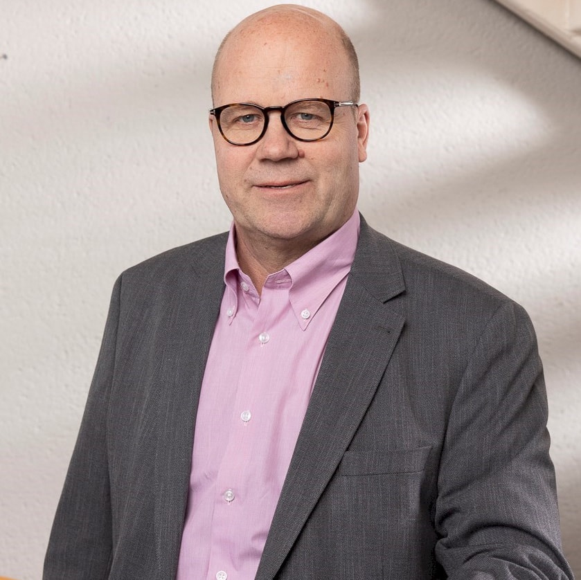 Jörgen Andersson En man i grå kostym och rosa skjorta står självsäkert i ett väl upplyst rum.
