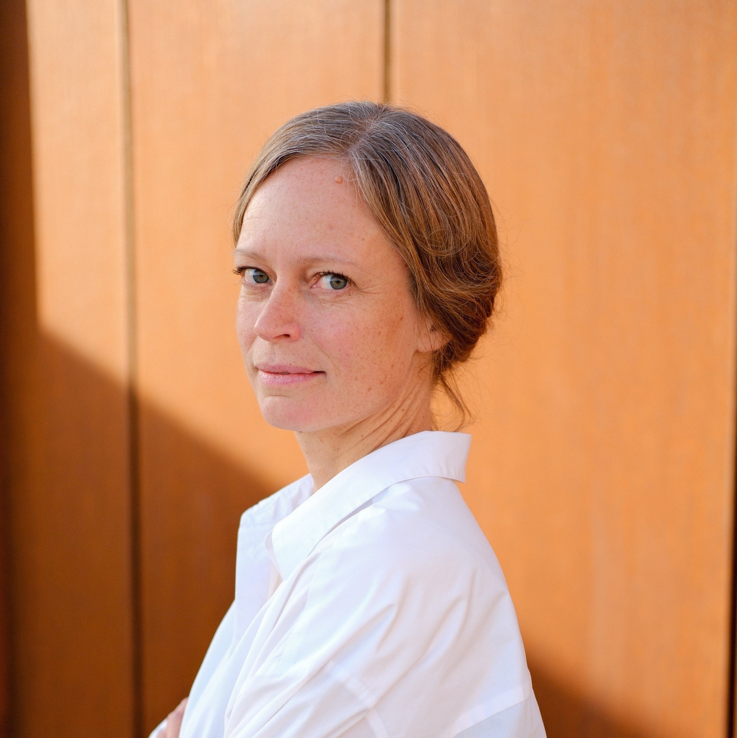 Kajsa Hedskog Kvinna med uppsatt hår i vit skjorta mot bakgrund i trä.