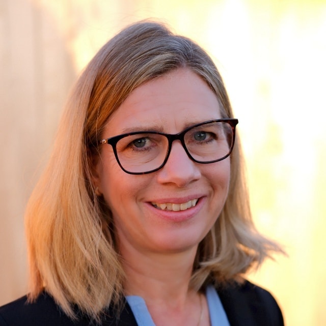 Leende person med blont hår och svarta glasögon.