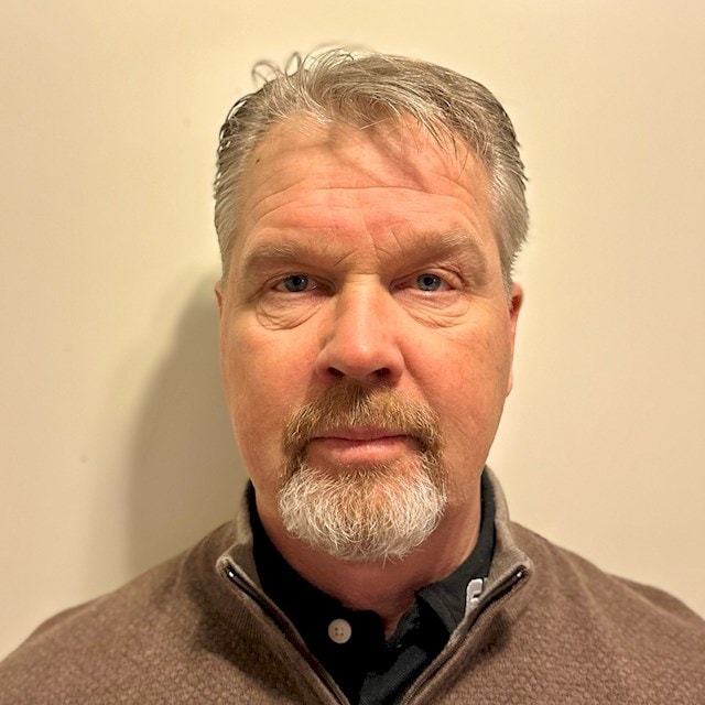 Kenneth Olsson En man med grått hår som bär en brun tröja med dragkedja över en svart skjorta står mot en beige vägg.