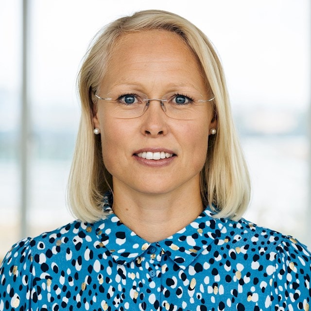 Maria Ronvall Kvinna med kort blont hår som bär en blå mönstrad blus med pärlörhängen.