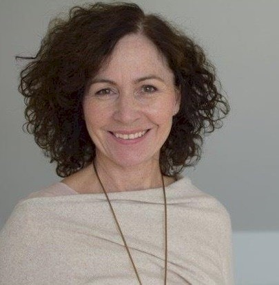 Woman with curly hair wearing a beige shawl and a long pendant against a gray background.