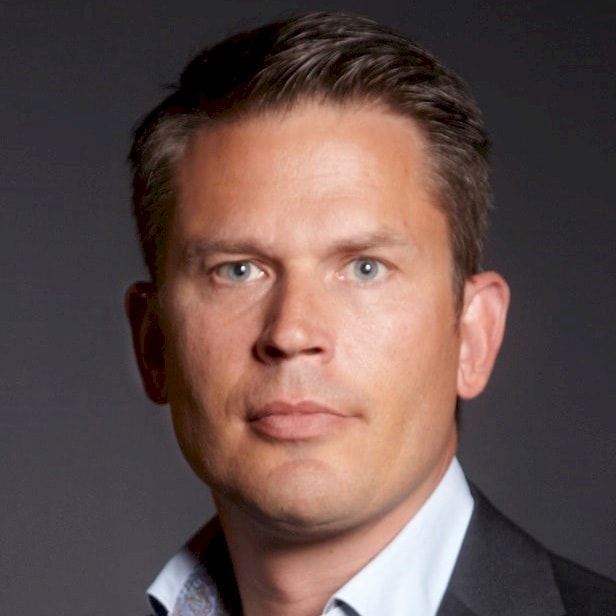 Nicklas Hagervall En man i svart kostym och vit skjorta med mönstrad krage mot en mörk bakgrund.