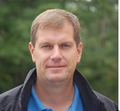 Peter Eltin Person i blå pikétröja och svart jacka står utomhus med grön suddig bakgrund.