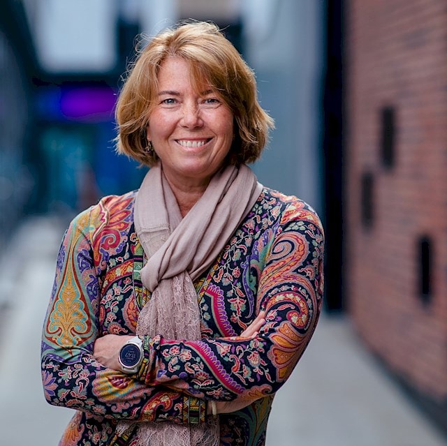 Synnøve Lyssand Sandberg Person wearing a colorful paisley dress and light pink scarf is standing outdoors.