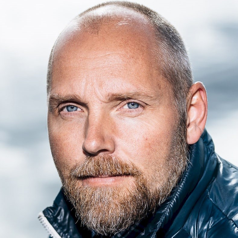 A person wearing a dark blue quilted jacket with a partially visible ear and grayish background.