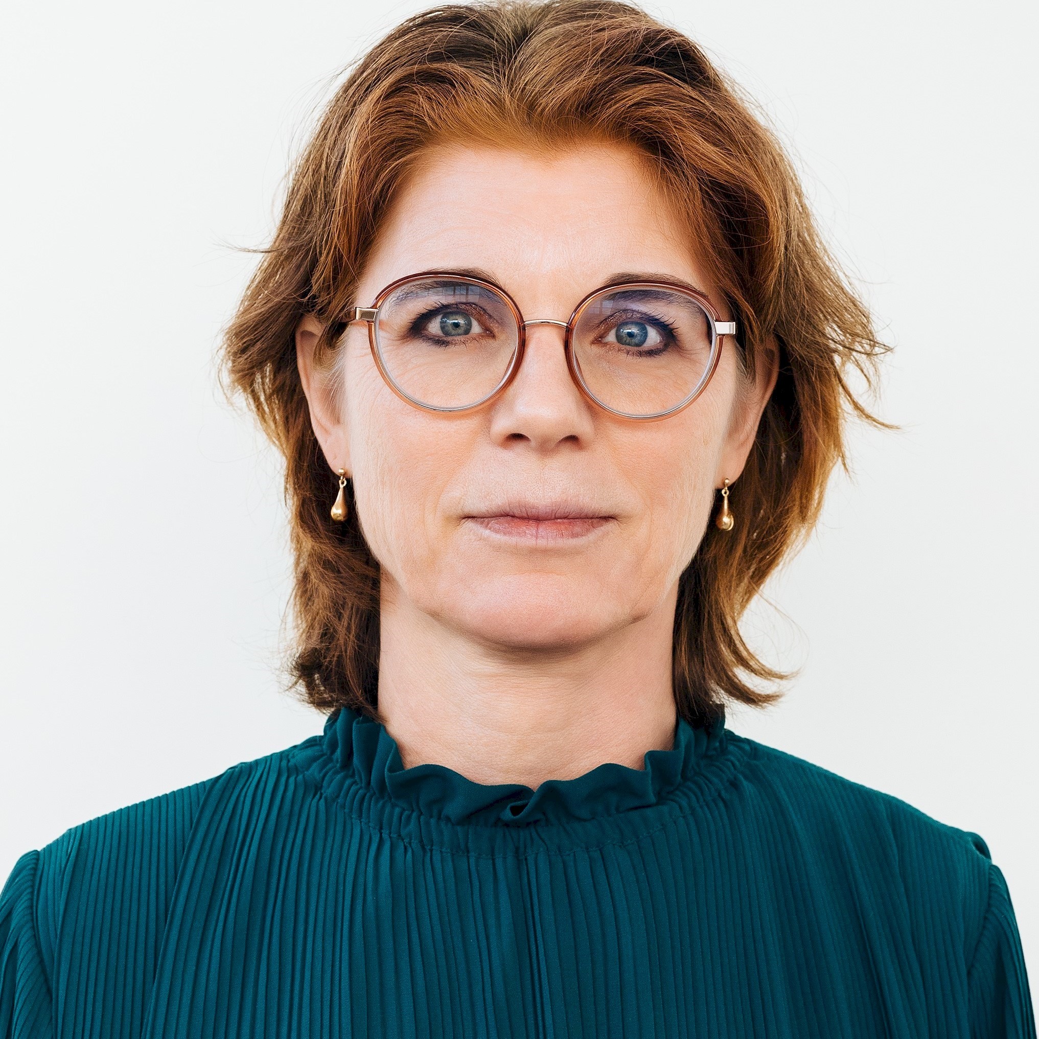 Ylva Gomér-Forss Person with short reddish hair wearing a green pleated blouse against a white background.