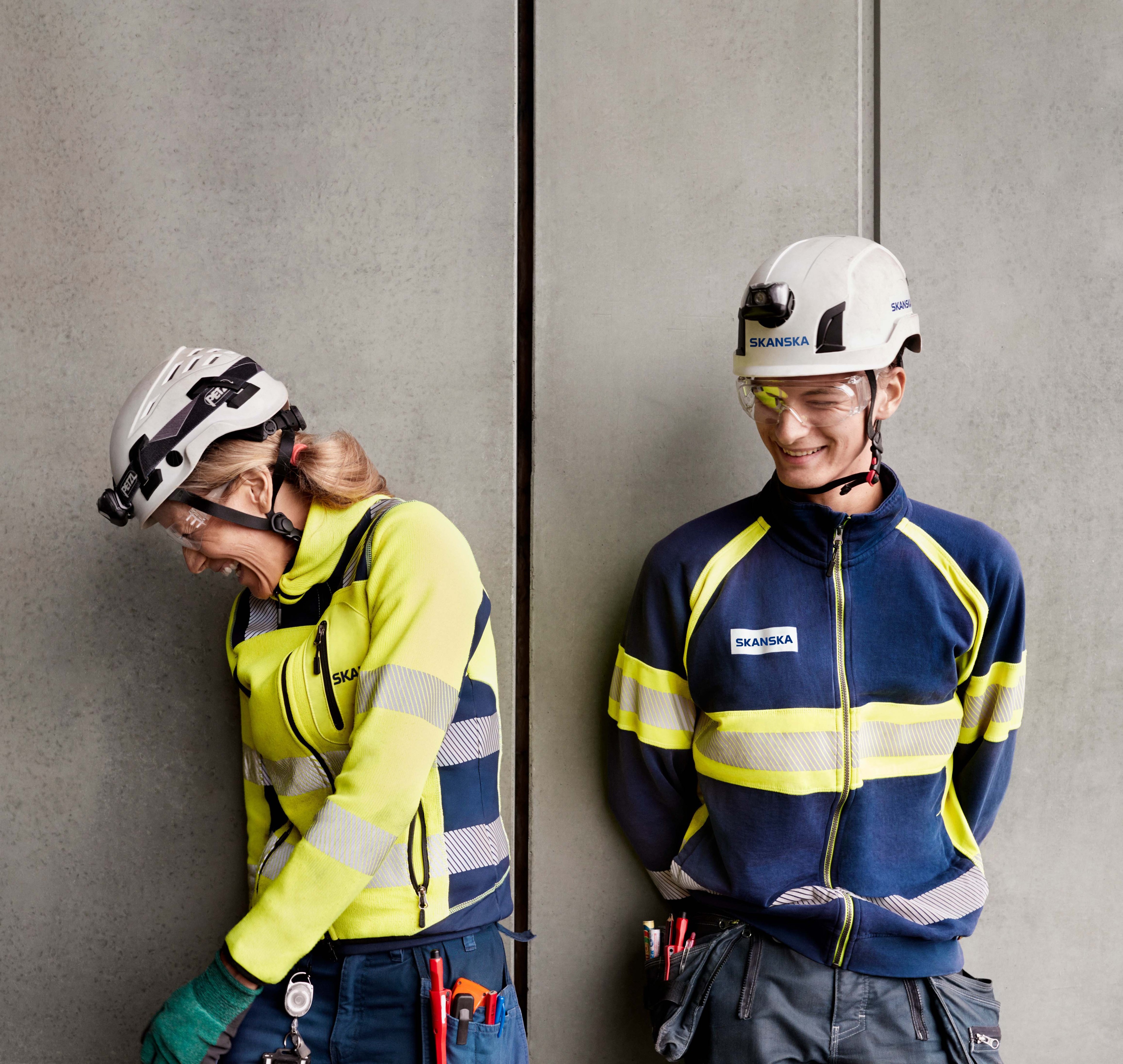 Två byggnadsarbetare i skyddsutrustning och hjälmar står mot en betongvägg.