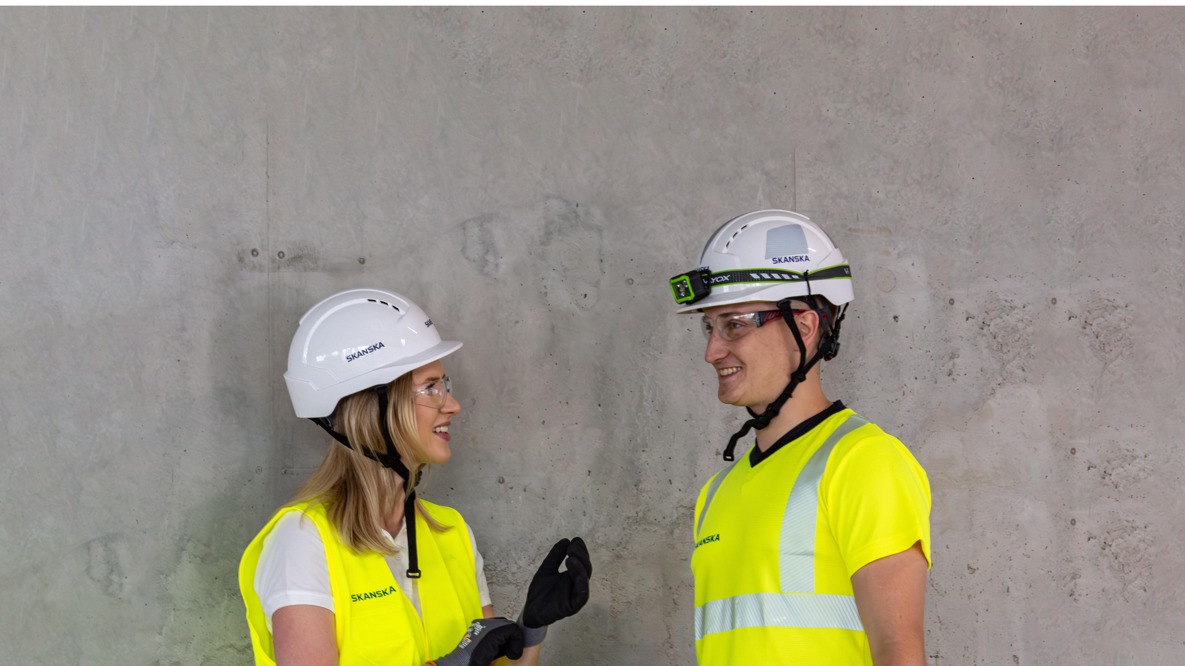 Två byggnadsarbetare med Skanska-hjälmar och varselvästar är engagerade i en diskussion.
