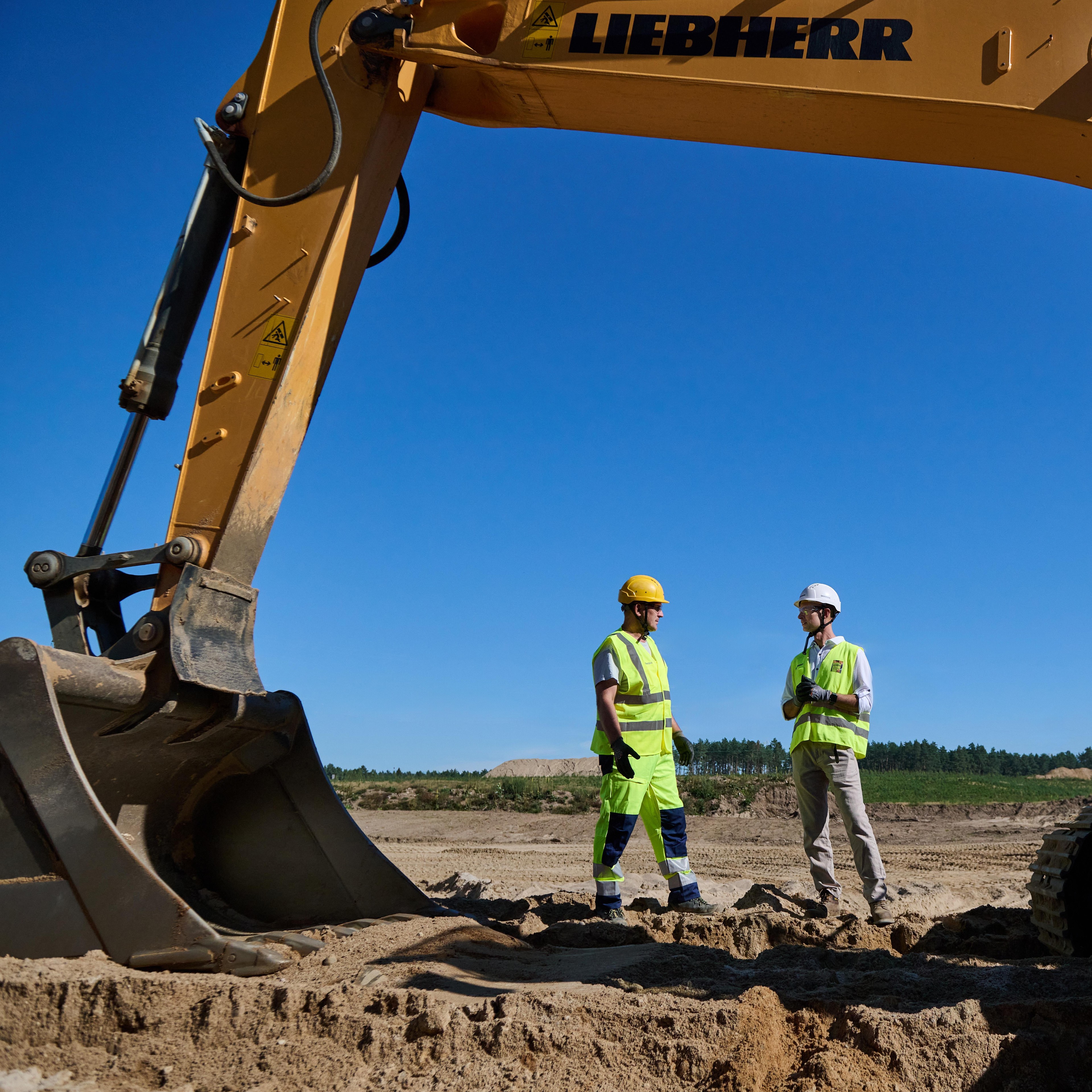 Two construction workers in safety equipment stand next to a large Liebherr excavator on a sandy site.