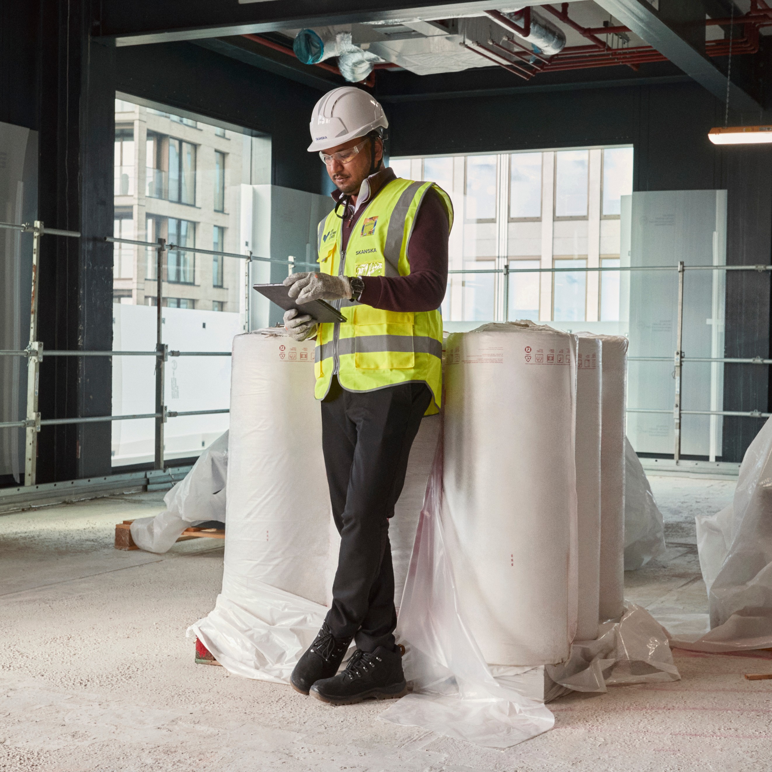 Placeholder Image with 3:2 Aspect Ratio for Design Use Construction worker in protective equipment, holding a tablet, standing by rolls of insulation material.
