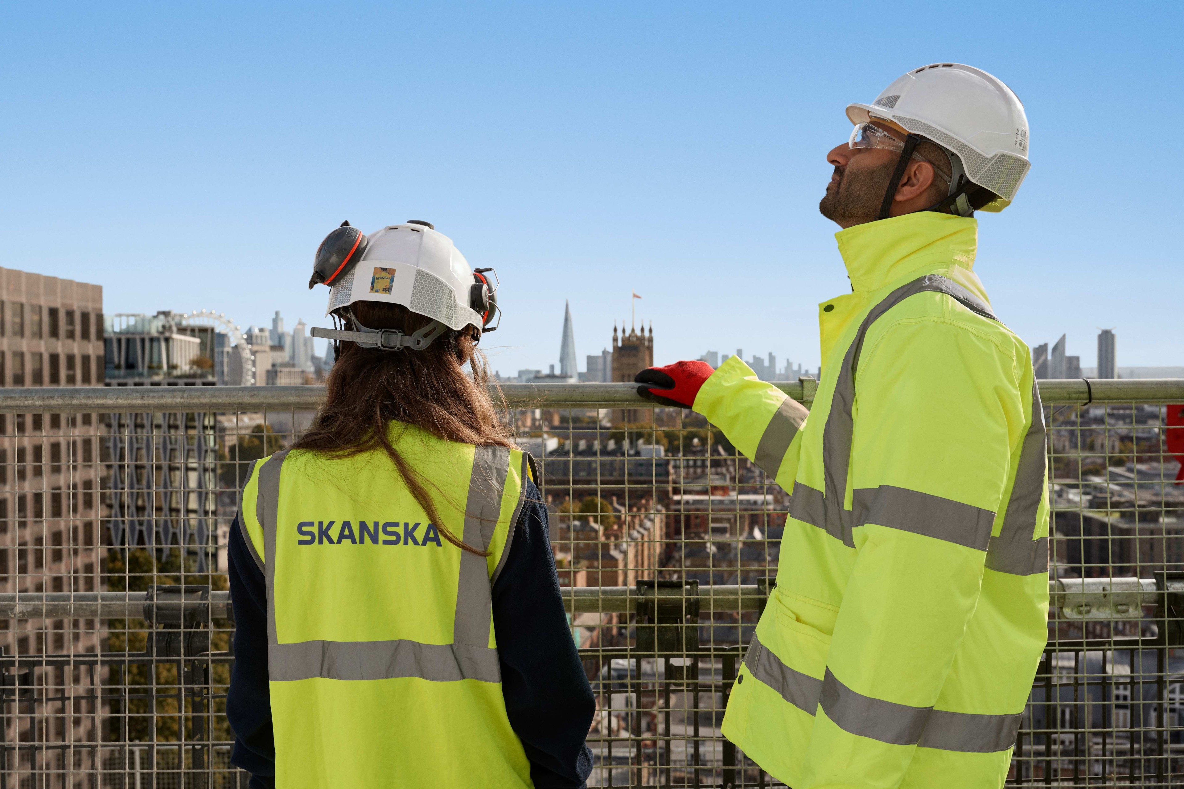 Två byggnadsarbetare i Skanska-utrustning blickar ut över en stadslandskap, bär hjälmar och säkerhetsvästar.