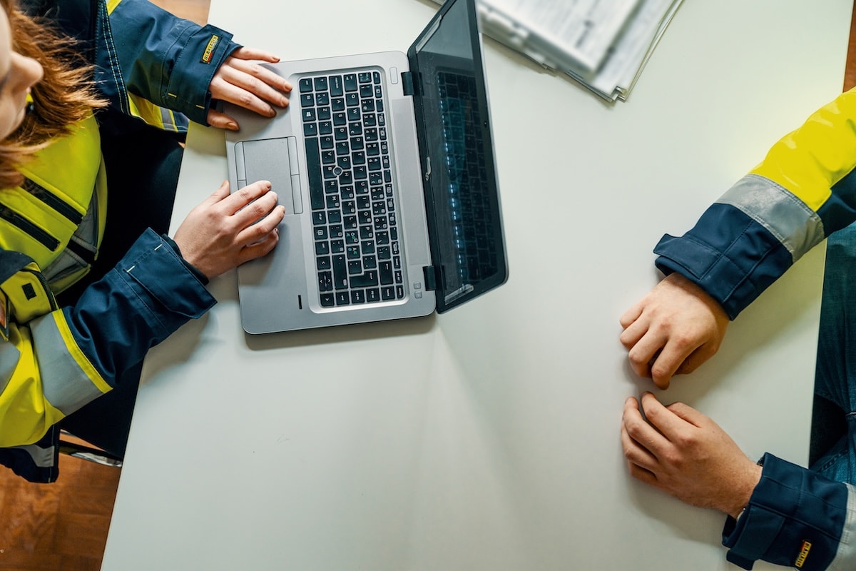 Two people in safety jackets are working at a laptop at a white table with documents nearby.