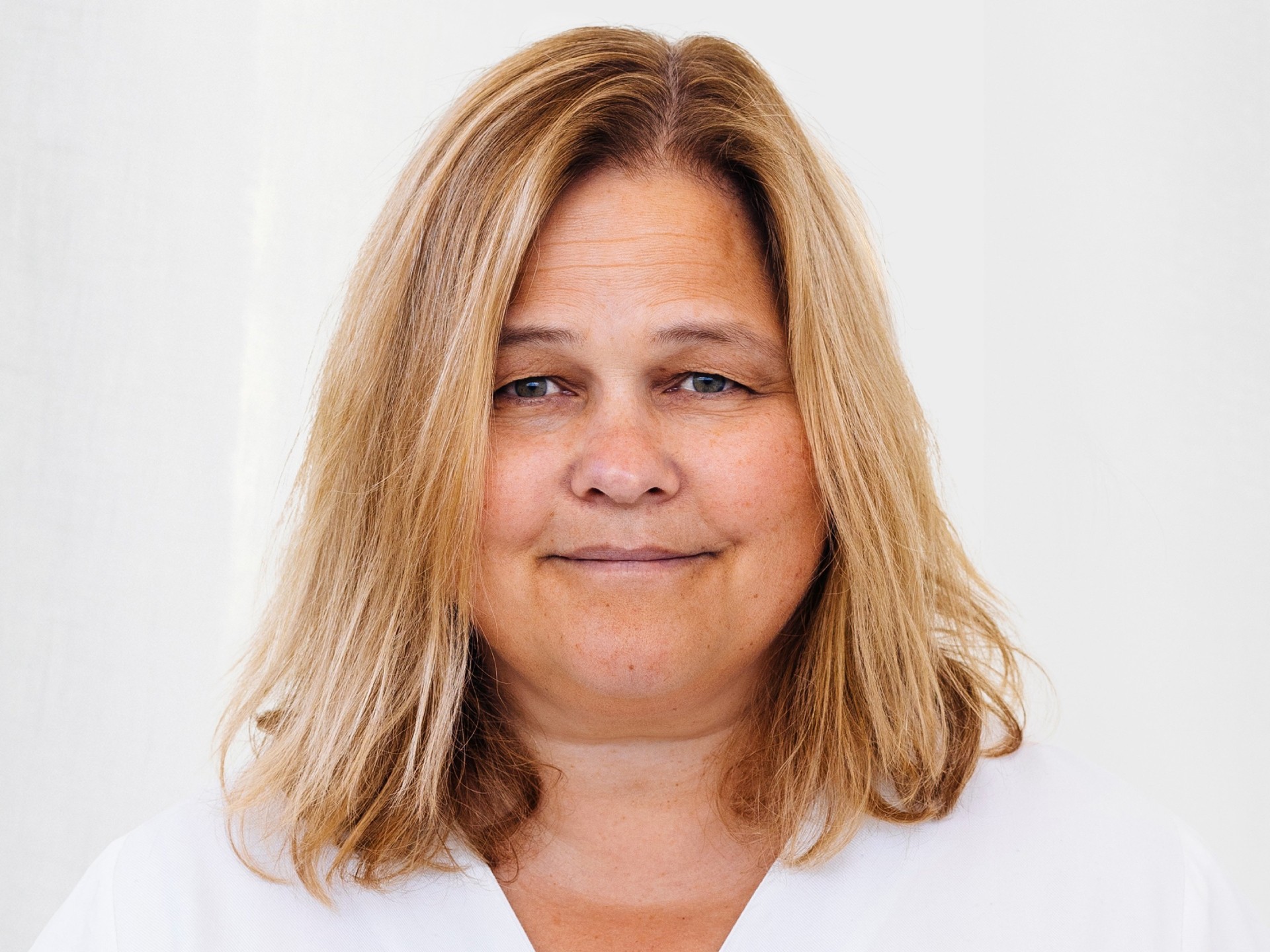 En person med axellångt blont hår som bär en vit topp mot en neutral bakgrund.