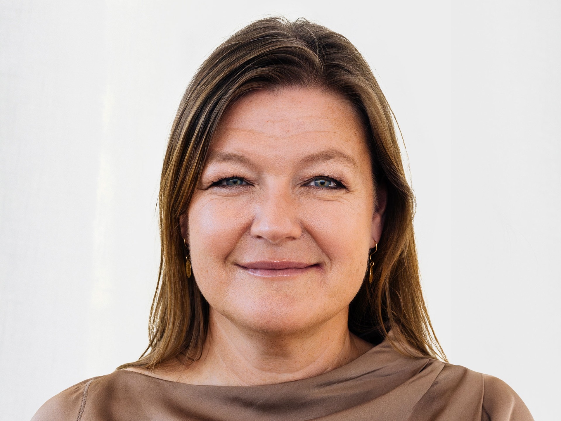 Kvinna med långt brunt hår som bär en taupe topp, mot en neutral bakgrund.