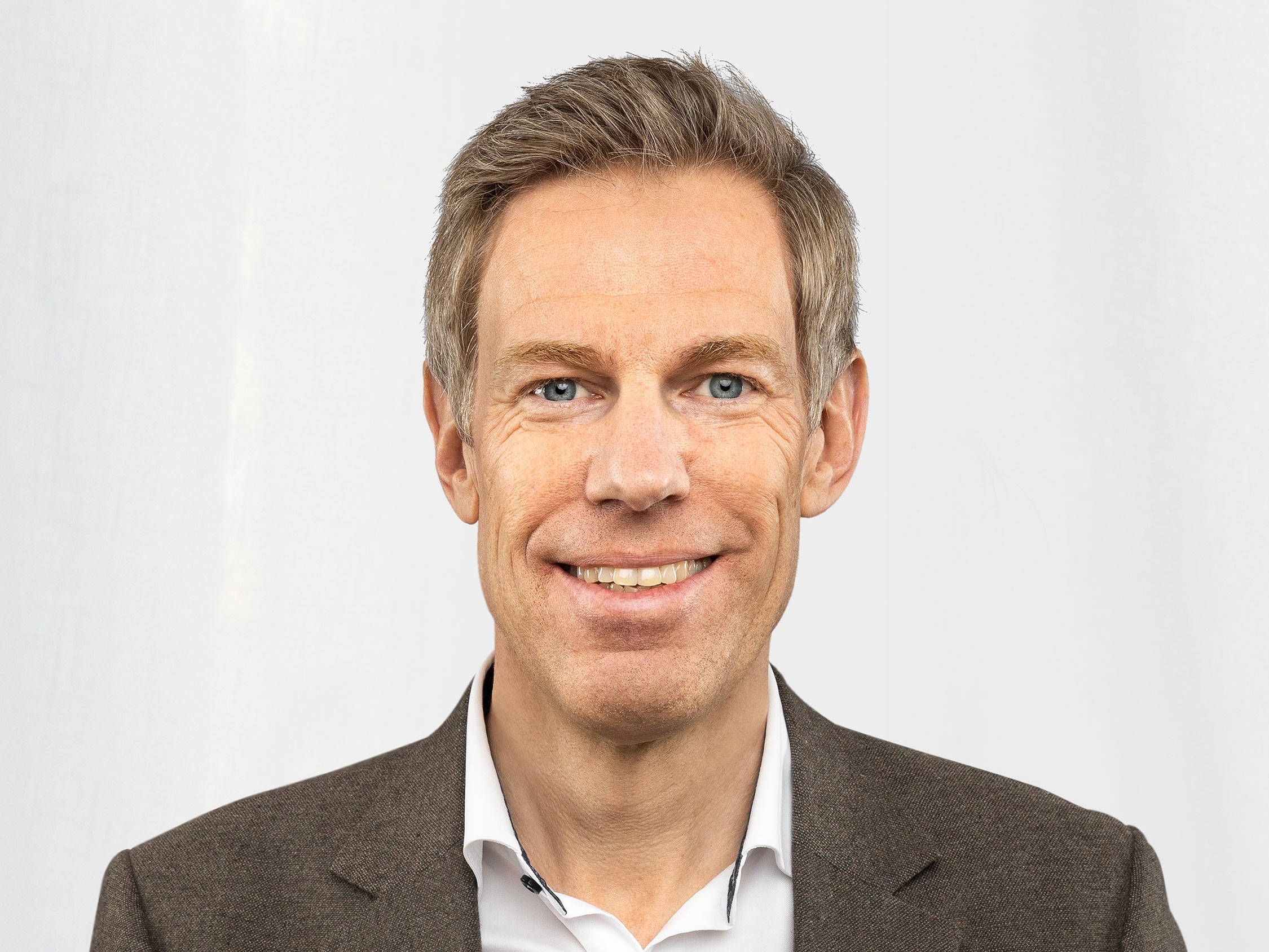 En person i brun kostym och vit skjorta med en mönstrad bröstnäsduk står mot en vit bakgrund.