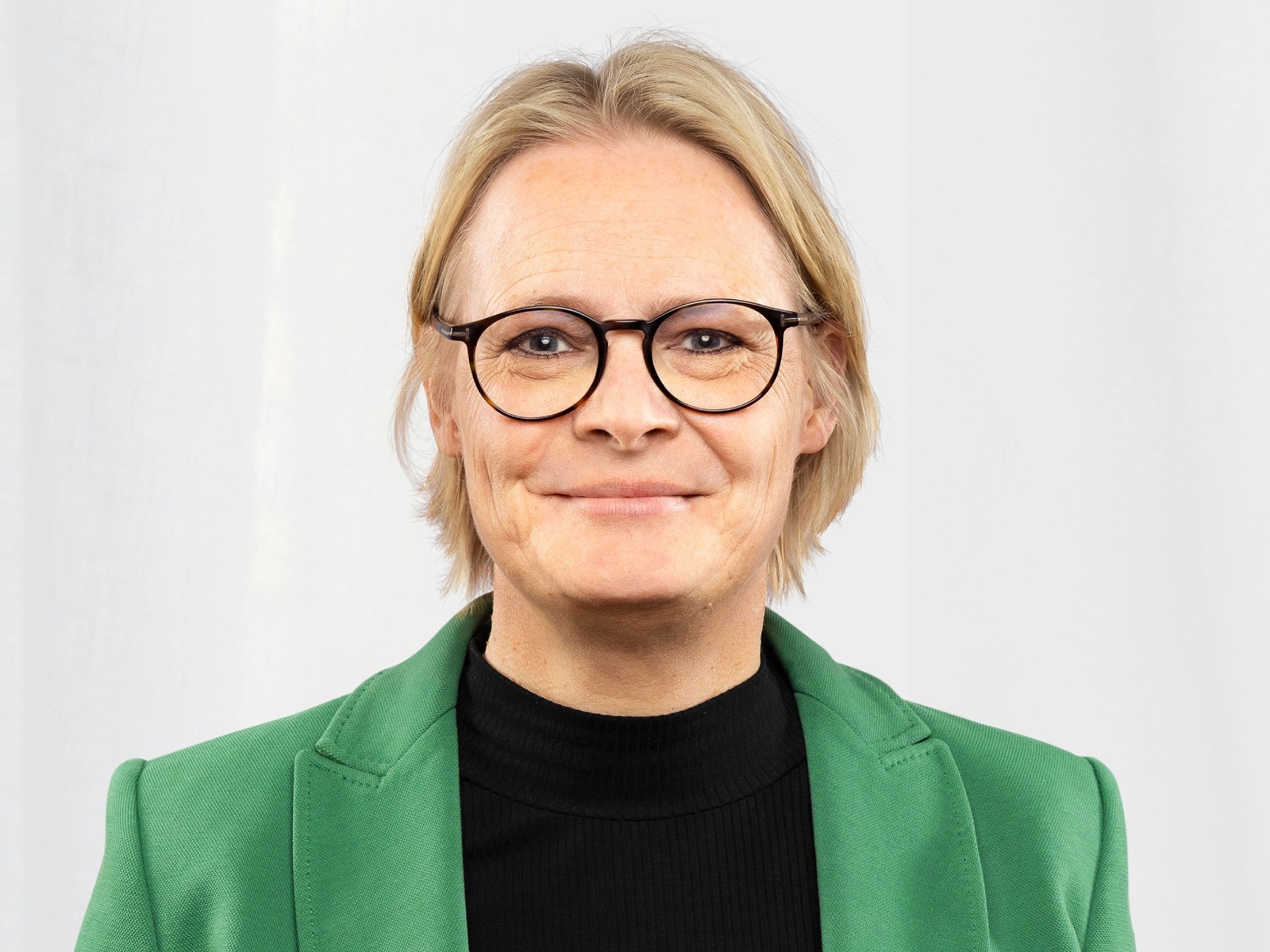 Person med blont hår som bär en grön kavaj och svart topp mot en enkel vit bakgrund.