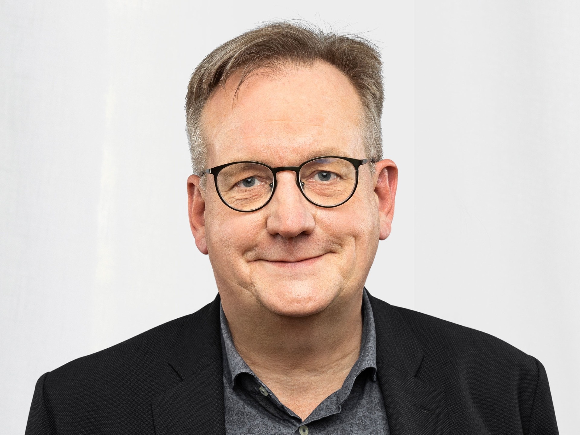 En man i en svart kostymjacka med en gråmönstrad skjorta mot en ljus bakgrund.